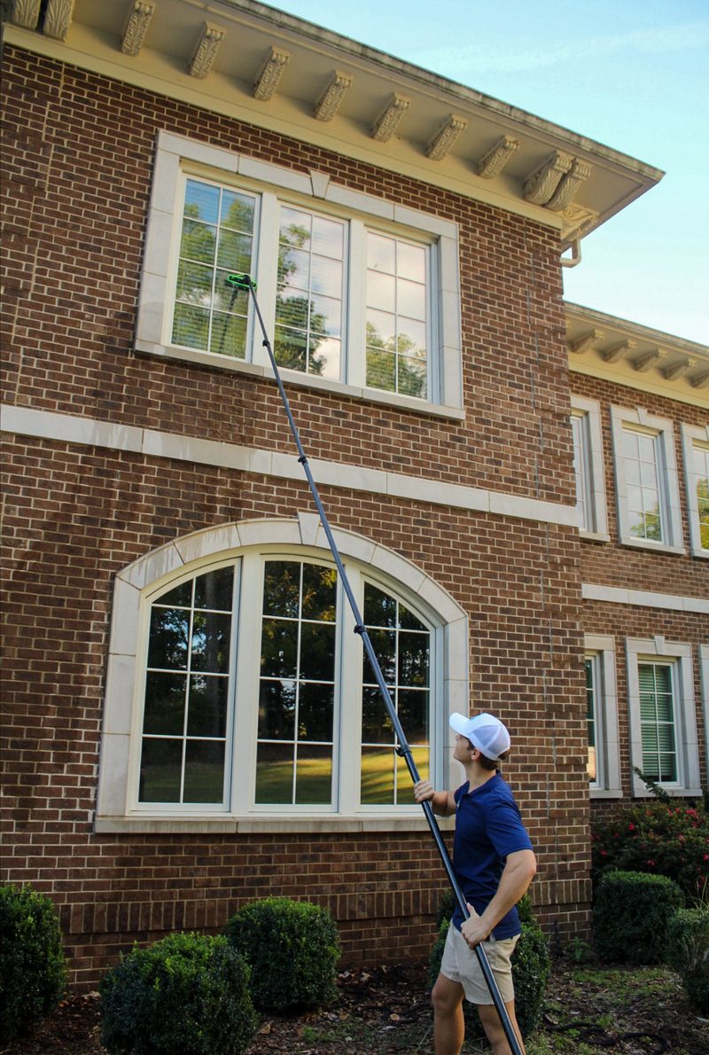 Technician in a navy polo and white cap cleaning a tall arched window on a brick home with a water-fed pole