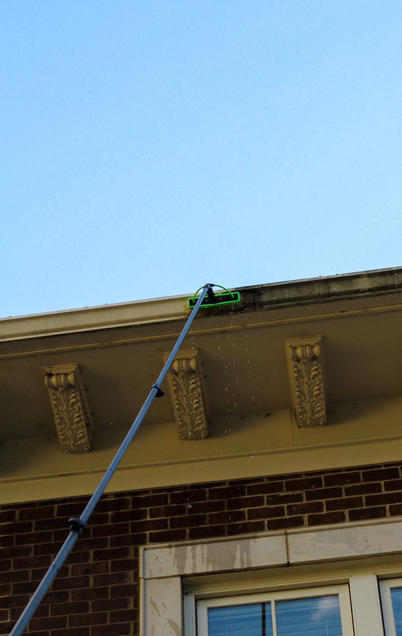 Water-fed pole brush reaching the roofline gutter of a brick home against a blue sky