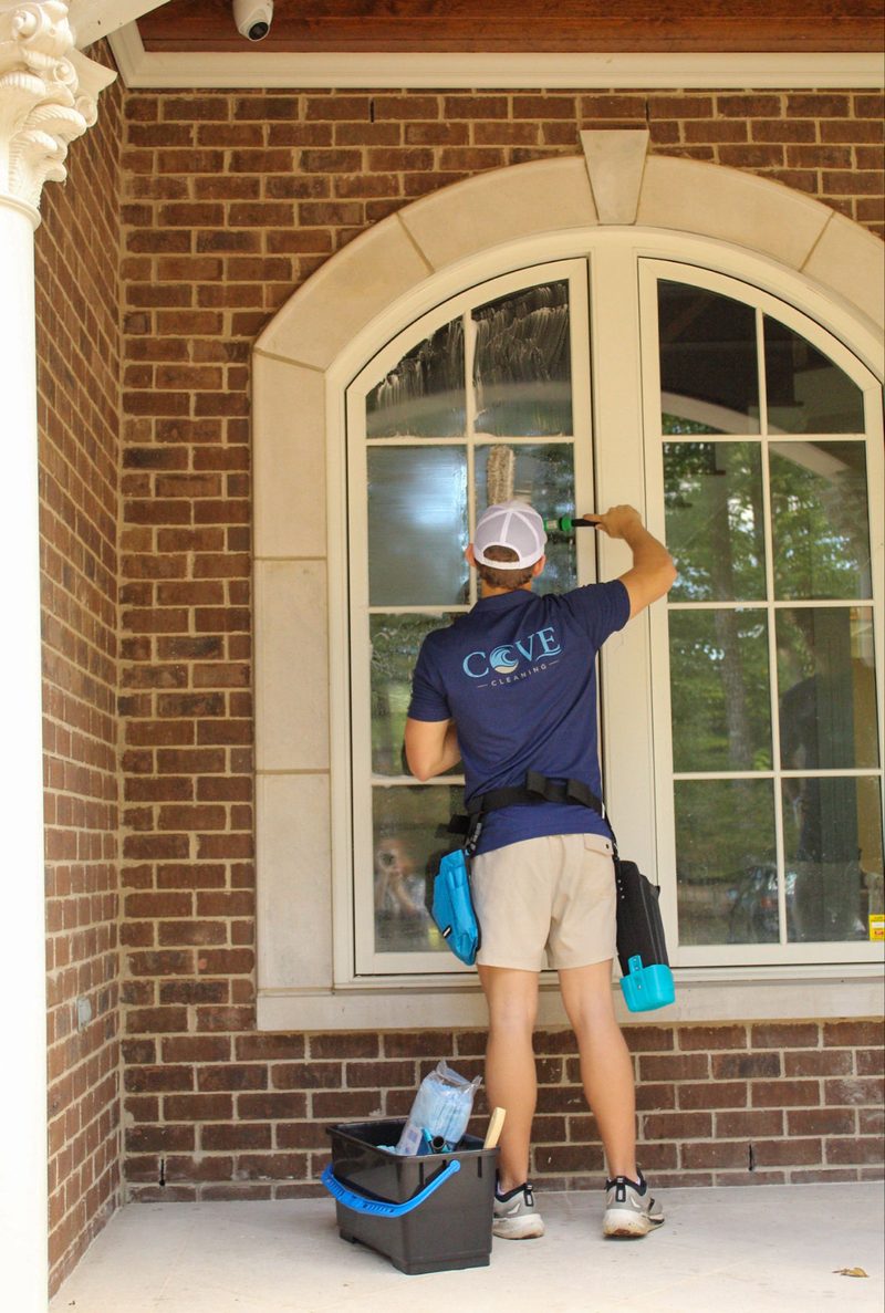 Cove Cleaning technician in a branded polo cleaning an arched window, with a bucket and tool belt